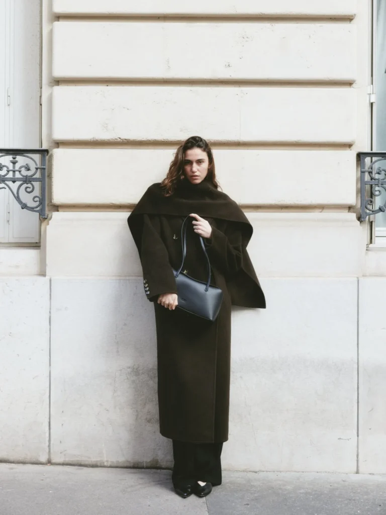 Woman in elegant dark coat holding a black handbag against a city wall.
