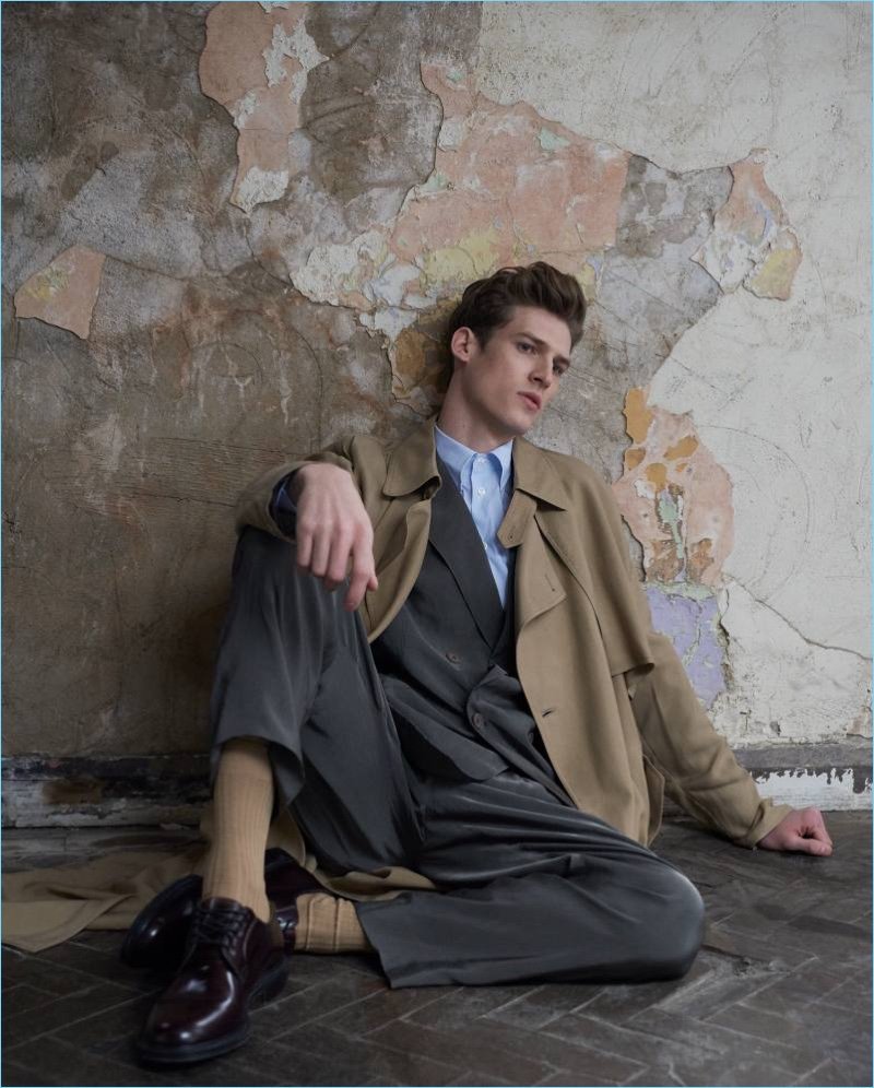Man in tailored trench coat and loafers posing against a textured wall