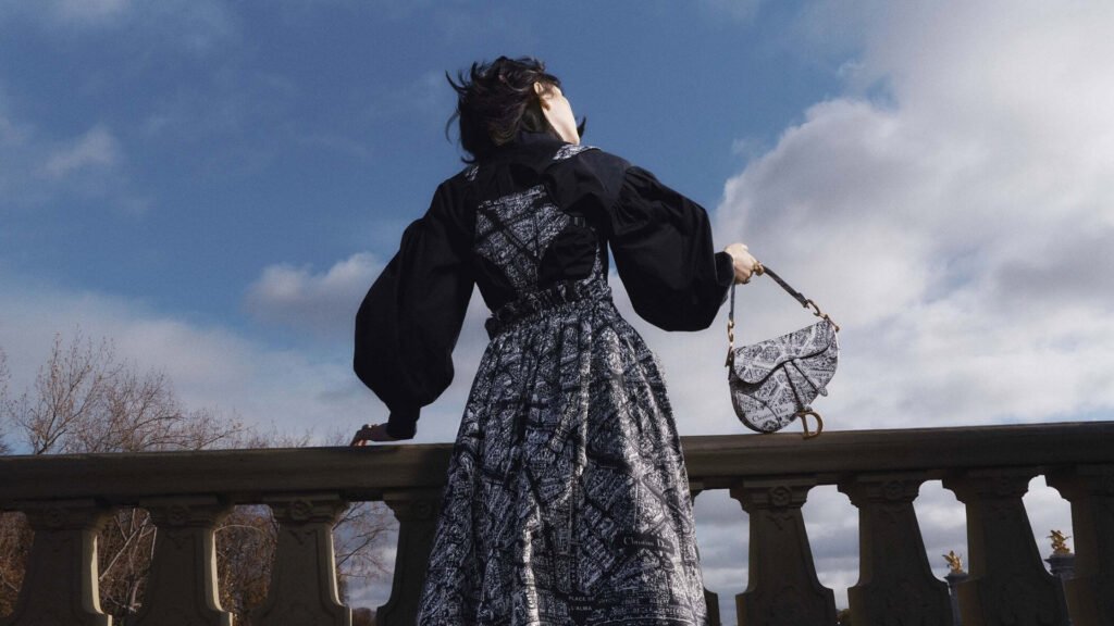 Model wearing a flowing Dior dress, holding a Dior saddle bag against a Parisian skyline.