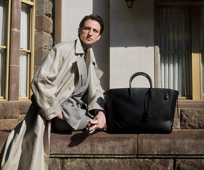 Model wearing a trench coat and carrying a large black Dunhill leather holdall.