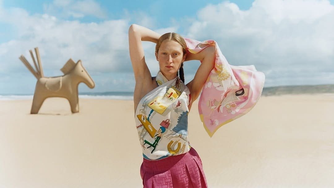 Model styling a large Hermès silk scarf with a pink skirt in a desert landscape.