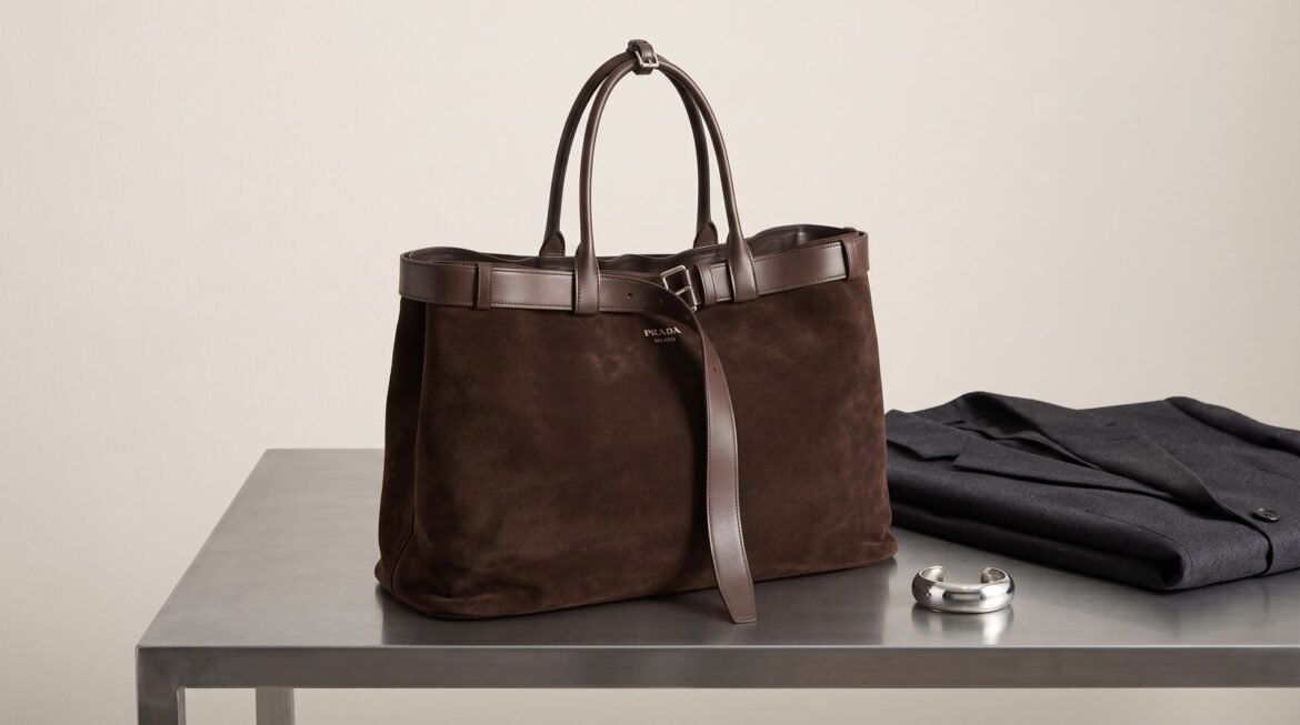 Dark brown suede Prada Buckle Bag on a brushed-steel table beside a charcoal blazer and silver cuff.