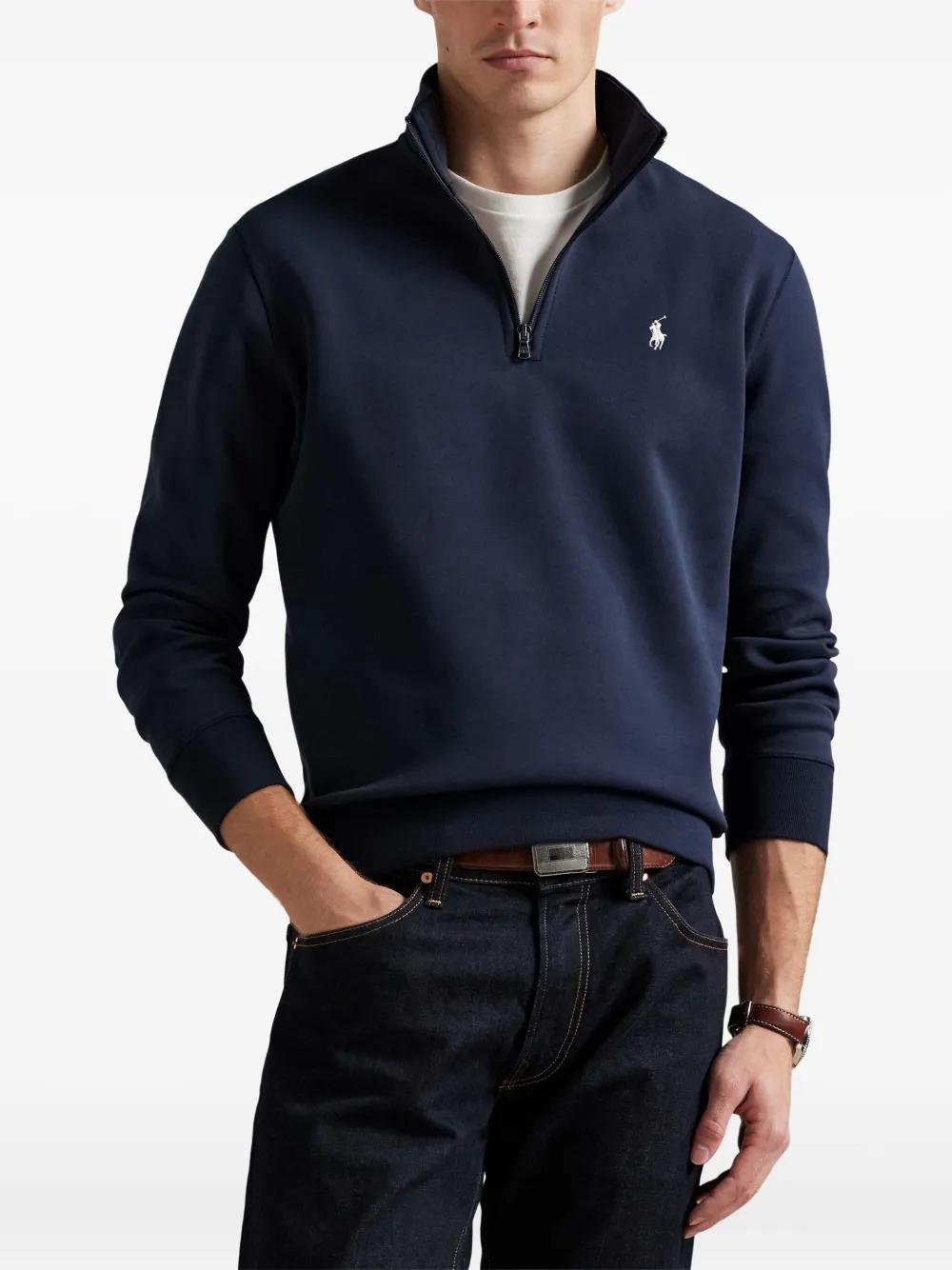 Model wearing a navy Polo Ralph Lauren quarter-zip with white trousers.