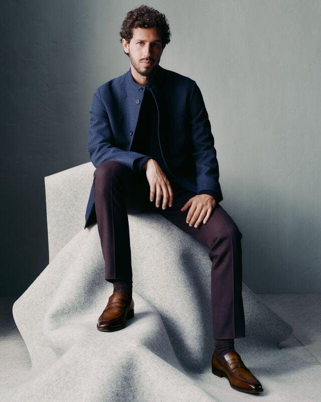 Seated model wearing brown Berluti shoes with a dark tailored suit.