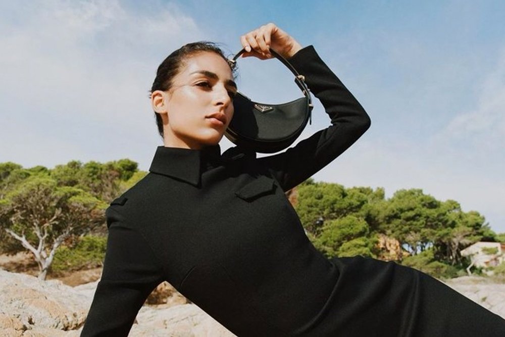 Model wearing a black dress and carrying a Prada Arqué shoulder bag outdoors.
