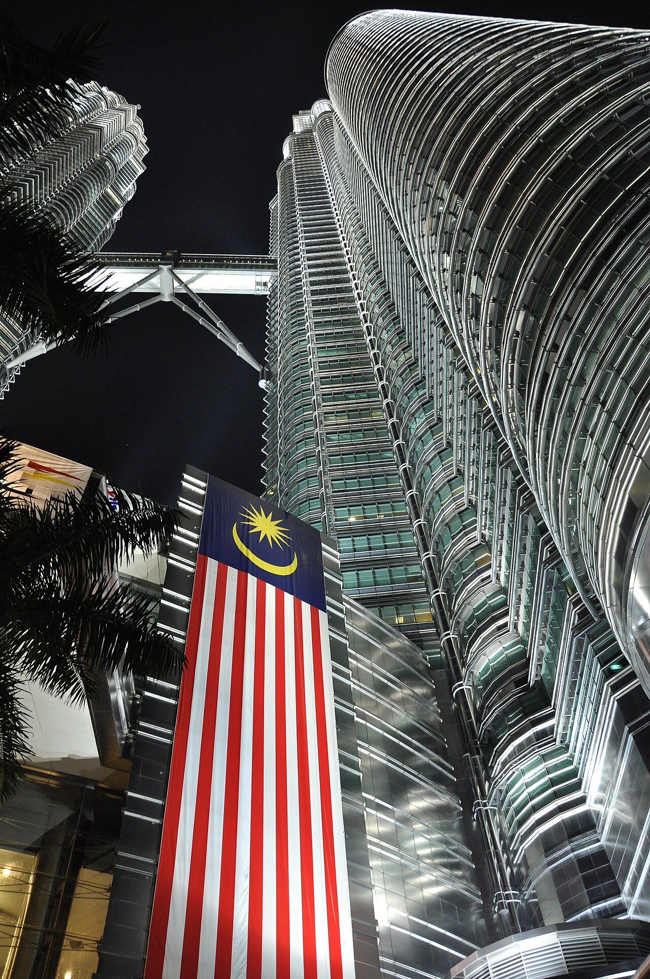 Upward view of the illuminated Petronas towers beside a Malaysian flag.